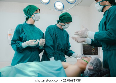 Group Of Young And Experienced Male And Female Surgeon With Nurse And Doctor Wearing Scrub, Mask And Headscarf Operating Young Woman In Operation Theatre In Modern Hospital