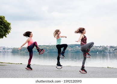 A group of young fit slim women in kangoo jumps, training in front of city lake in summer. Three sportswomen, wearing colorful sports outfit, doing exercises outside. Healthy lifestyle concept.
