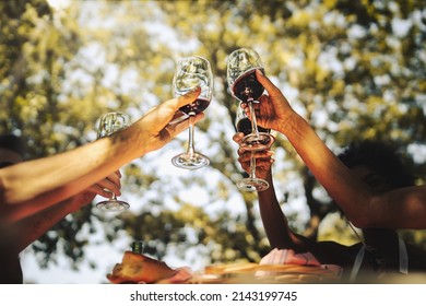Group of young friends celebrating friendship rising hands holding red wine glasses in the countryside at the picnic