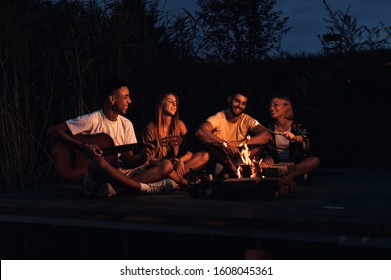 Group of young friends enjoying at the lake at night. They sitting around the fire singing and having fun at camping. - Powered by Shutterstock