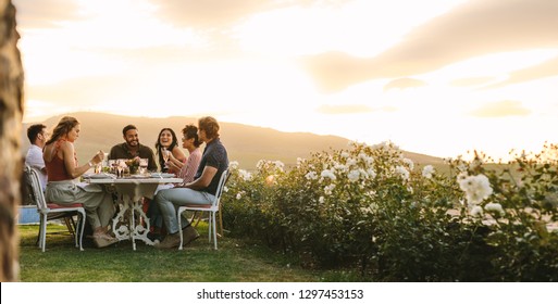 Group Of Young Friends Hanging Out With Drinks At Outdoors Dinner Party. Young Men And Women Sitting Around A Table Having Food And Drinks.