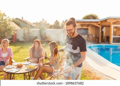 Group Of Young Friends Having A Backyard Barbecue Party By The Swimming Pool, Man Grilling Meat And Drinking Beer In The Foreground While Women Are Sitting And Relaxing In The Background