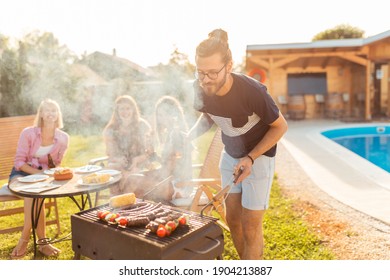 Group of young friends having a backyard barbecue party by the swimming pool, man grilling meat and drinking beer in the foreground while women are sitting and relaxing in the background - Powered by Shutterstock