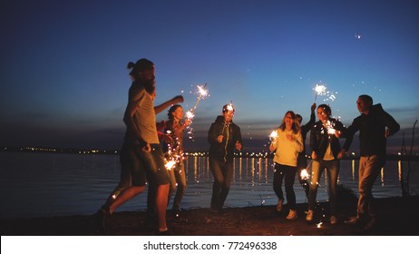 Group Of Young Friends Having A Beach Party. Friends Dancing And Celebrating With Sparklers In Twilight Sunset