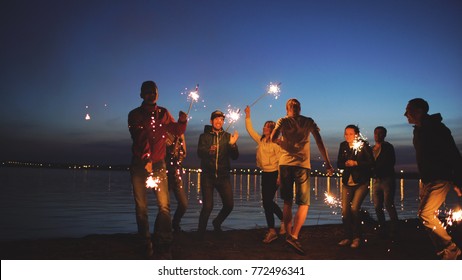 Group Of Young Friends Having A Beach Party. Friends Dancing And Celebrating With Sparklers In Twilight Sunset