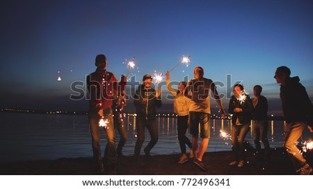 Group of young friends having a beach party. Friends dancing and celebrating with sparklers in twilight sunset
