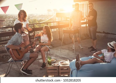 Group of young friends having fun at rooftop party, making barbecue, drinking beer and enjoying hot summer days. Focus on the girl sitting on the chair - Powered by Shutterstock