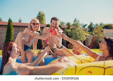 Group of young friends having fun at summertime swimming pool party, making a toast with watermelon popsicles while sunbathing by the pool - Powered by Shutterstock