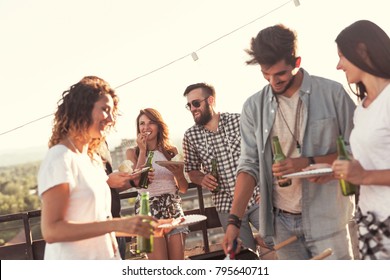 Group Of Young Friends Having Fun At Rooftop Party, Making Barbecue And Enjoying Hot Summer Days. Focus On The Couple In The Background