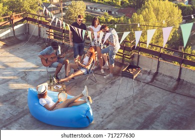 Group Of Young Friends Having A Rooftop Party, Playing The Guitar And Enjoying Hot Summer Days