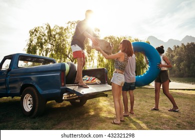 Group of young friends on picnic by the lake. Men and women unloading pickup truck on camping trip, carrying picnic basket and inflatable tube on sunny day. - Powered by Shutterstock