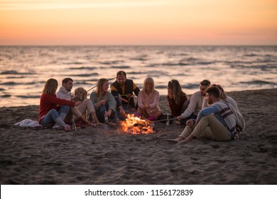 Group Of Young Friends Sitting By The Fire At Autumn Beach, Grilling Sausages And Drinking Beer, Talking And Having Fun