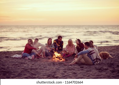 Group of young friends sitting by the fire at autumn beach, grilling sausages and drinking beer, talking and having fun filter - Powered by Shutterstock