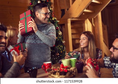 Group Of Young Friends Sitting Next To A Nicely Decorated Christmas Tree, Exchanging Christmas Presents And Having Fun