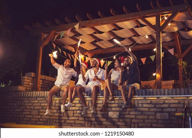 Group of young friends sitting on a poolside brick wall, having fun on a New Year's Eve, dancing, drinking beer and waving with sparklers at midnight - Powered by Shutterstock