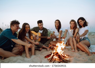 Group of young friends sitting on beach and fry sausages. One man is playing guitar. Summer holidays, vacation, relax and lifestyle consept. Camping time. - Powered by Shutterstock