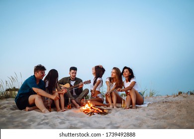 Group of young friends sitting on beach and fry sausages. One man is playing guitar. Summer holidays, vacation, relax and lifestyle concept. Camping time. - Powered by Shutterstock