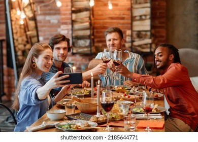 Group Of Young Friends Taking Selfie Photo At Table During Dinner Party And Clinking Glasses