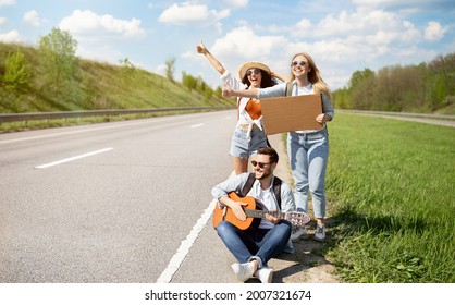 Group of young friends thumbing their ride on road with empty sign, playing guitar, having autostop journey, free space. Millennial hitchhikers having fun road trip, enjoying summer adventure