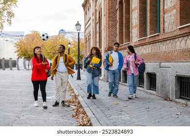 group of young friends walking along a city street, with one person playfully tossing a soccer ball in the air. - Powered by Shutterstock