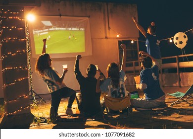 Group of young friends watching a football match on a building rooftop, drinking beer and cheering. Selective focus on the people in the middle - Powered by Shutterstock