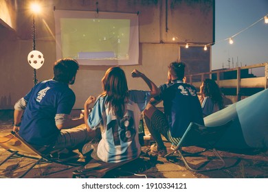 Group of young friends watching a football match on a building rooftop terrace, drinking beer and cheering for their team