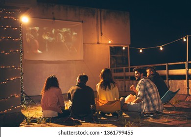 Gruppe junger Freunde, die einen Film auf einer Dachterrasse im Gebäude sehen, Popcorn essen, Bier trinken und Spaß haben