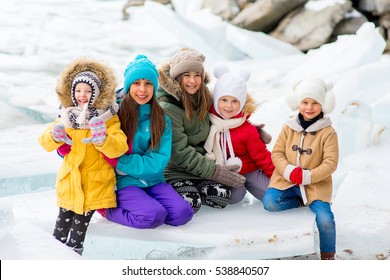 Group Of Young Girls Sitting At The Ice Block On The Frozen Lake. Outdoors