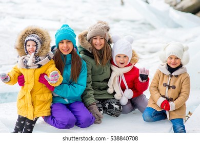 Group Of Young Girls Sitting At The Ice Block On The Frozen Lake. Outdoors