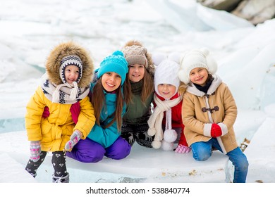 Group Of Young Girls Sitting At The Ice Block On The Frozen Lake. Outdoors