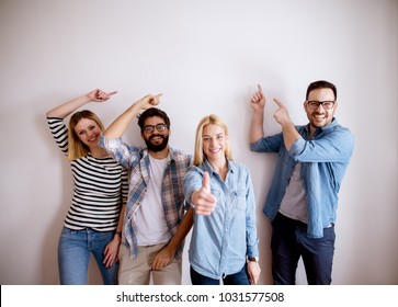 Group Of Young Handsome Colleague People Standing Against The Wall Showing At Empty Editable Space Above While One Girl Standing With Thumb Up And Looking At The Camera.