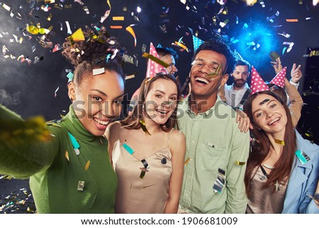 Group of young happy multiracial people wearing birthday hats making a selfie on mobile phone, confetti falling in the air