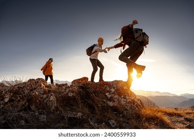 Group of young hikers with backpacks walks with backpacks and helps each other in climbing in sunset mountains