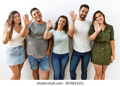 Group of young hispanic friends standing together over isolated background waiving saying hello happy and smiling, friendly welcome gesture 
