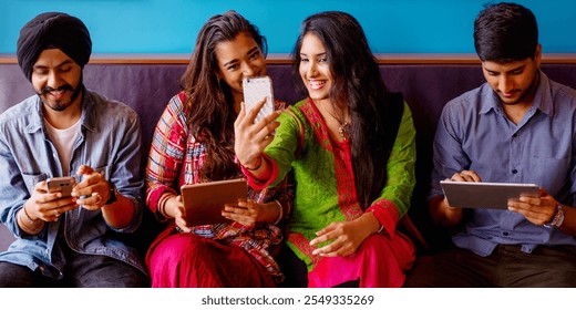 Group of young Indian friends sitting together, using smartphones and tablets. Diverse group enjoying technology, taking selfies, sharing moments on devices. Indian young friends using digital devices - Powered by Shutterstock