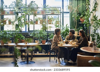 Group of young intercultural friends sitting by table in the corner of cafe with variety of green plants, having chat and coffee with snacks