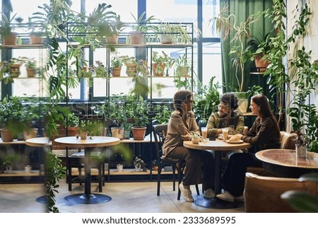 Group of young intercultural friends sitting by table in the corner of cafe with variety of green plants, having chat and coffee with snacks