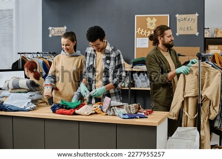 Group of young intercultural volunteers sorting second hand clothes on table and choosing casualwear on rack for people in need