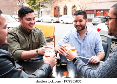 Group Of Young Latino Friends, Sitting Outside A Bar On An Autumn Afternoon, Talking To Each Other And Smiling, Making A Toast With Craft Beer, Bar And Lifestyle Concept.