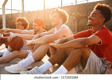 Group of young laughing multiethnic men basketball players resting at the sport ground, talking