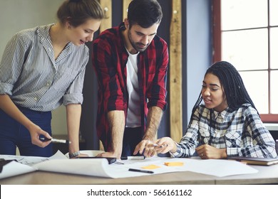 Group of young male and female students sharing ideas while working on project correcting mistakes in architectural drawings without using technologies collaborating in coworking space with bid desk