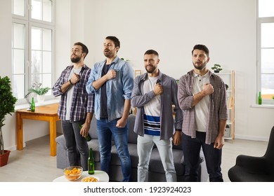 Group of young men supporting favourite football team. Happy proud soccer fans standing together in living-room, holding hand over heart and singing national country anthem before watching match on TV