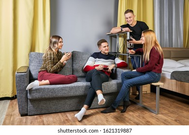 A Group Of Young Men And Women Drink Alcohol At A College Dorm Party. Drunk On The University Campus, Undergraduates Are Sitting In The Bedroom With Glasses Of Wine In Their Hands.