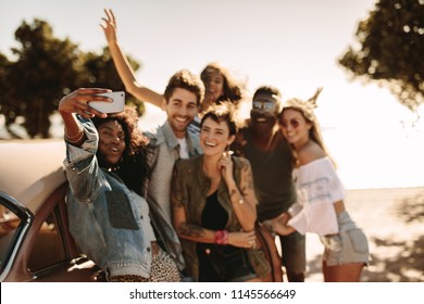 Group Of Young Men And Women Enjoying On Road Trip Standing By The Car And Taking Selfie. Multiracial Group Of Friends Having Fun Outdoors Taking Selfie With Smart Phone.