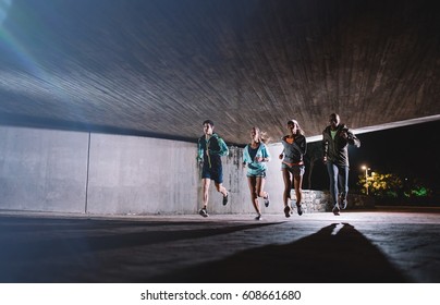 Group Of Young Men And Women Running Together At Night. Healthy Young People Training Together Under A Bridge In City.