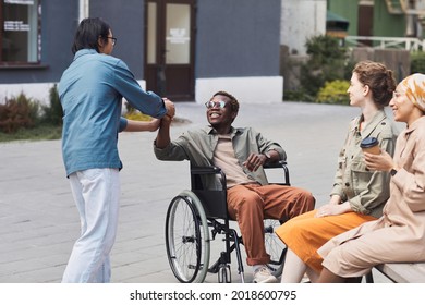 Group of young multi-ethnic friends including disabled man in wheelchair gathering together outdoors, men making handshake