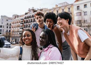 A group of young multiethnic people are posing for a picture, with one of them taking a photo of the group. Scene is happy and friendly, as the group is smiling and enjoying each other's company