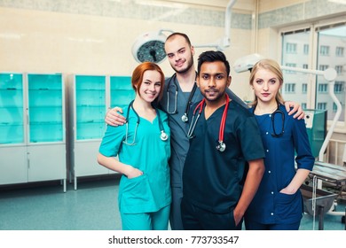 Group Young Multiracial Medical Students With Surgeons Hugging Each Other Looking With A Smile At The Camera. Hospital Operating Room In The Background