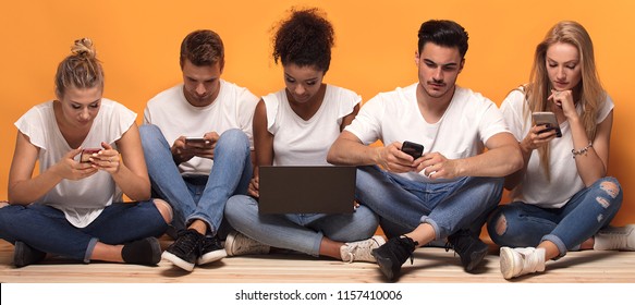 Group Of Young Multiracial People Looking At Mobile Phones, Sitting On The Floor, Spending Time Together.