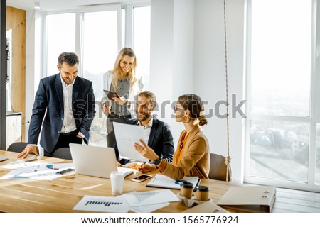 Group of a young office employees dressed casually in the suits having some office work at the large meeting table in the bright sunny room